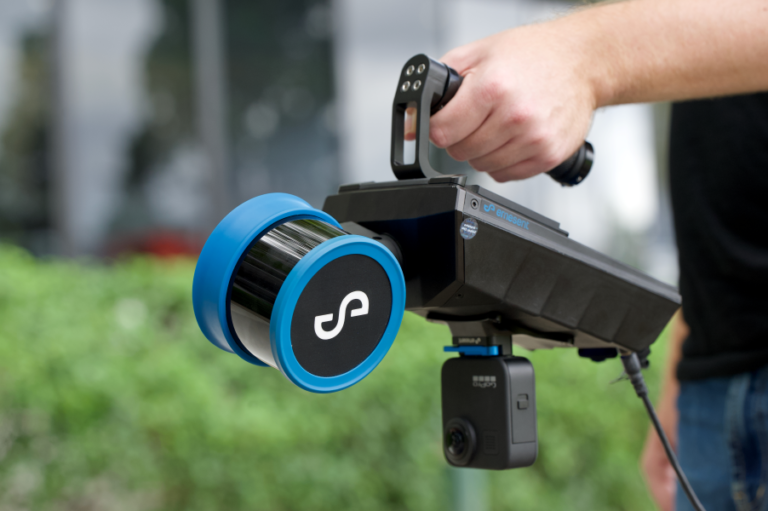 Person hält im Freien einen schwarz-blauen tragbaren 3D-Scanner mit angeschlossener Kamera.