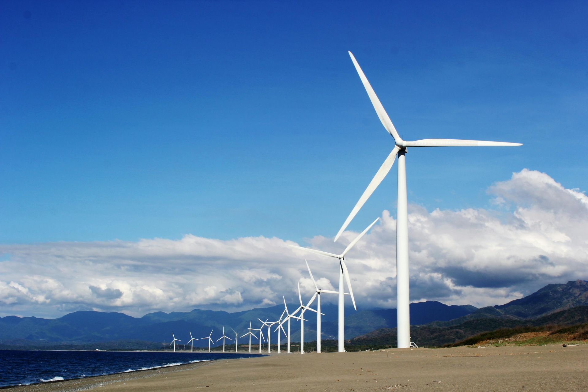 A row of wind turbines on a beach with mountains in the background