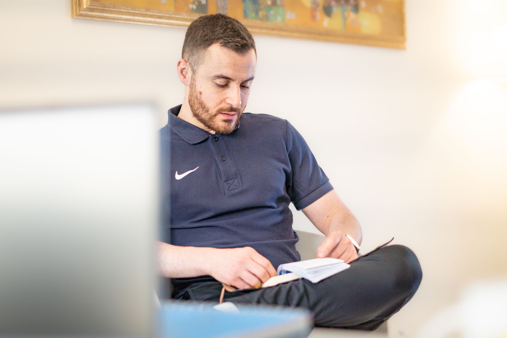 Uomo con polo blu navy, a gambe incrociate, che scrive su un quaderno. All'interno, computer portatile in primo piano, dipinto sul muro.