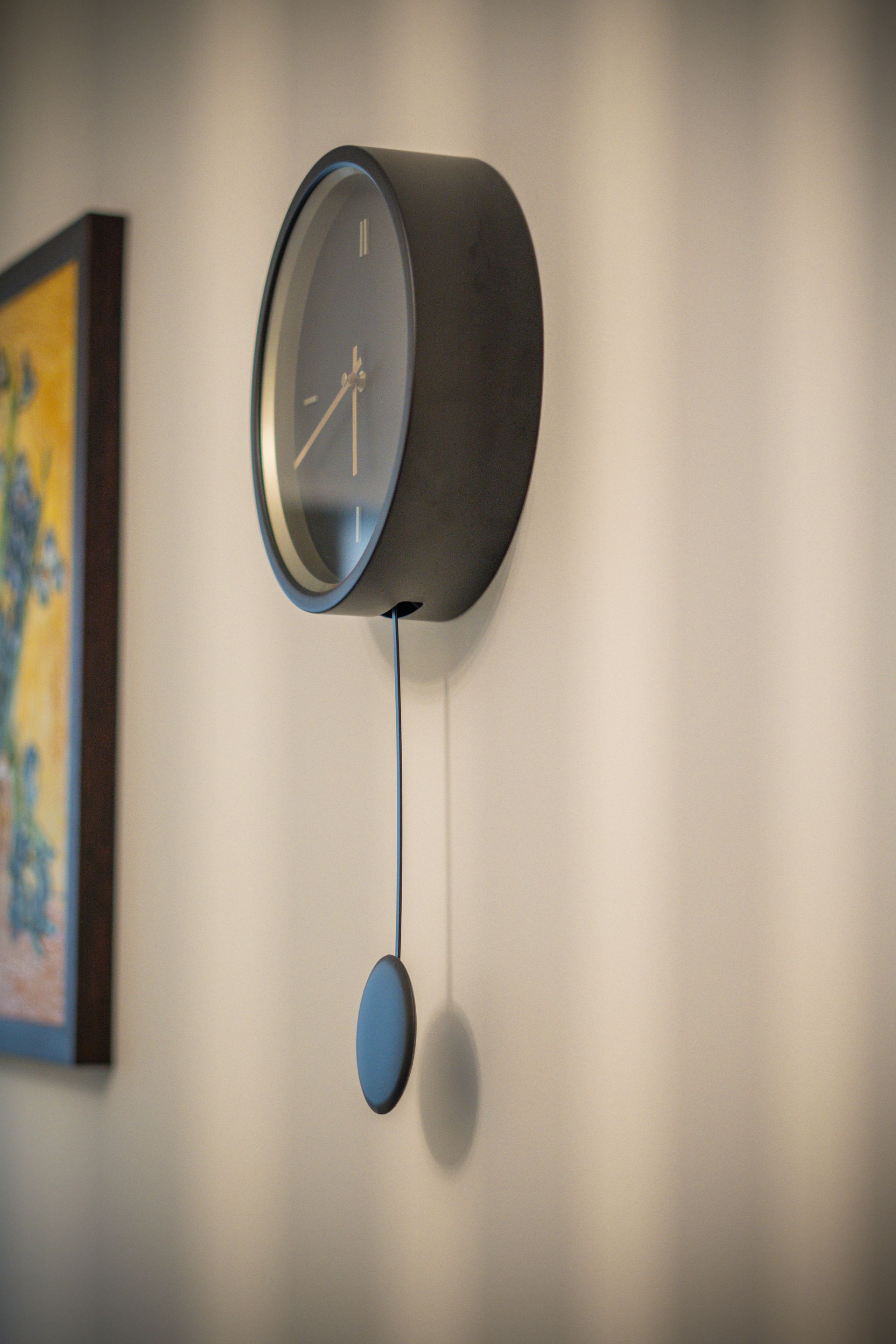 Orologio da parete nero con pendolo, appeso a una parete beige accanto a un dipinto incorniciato.