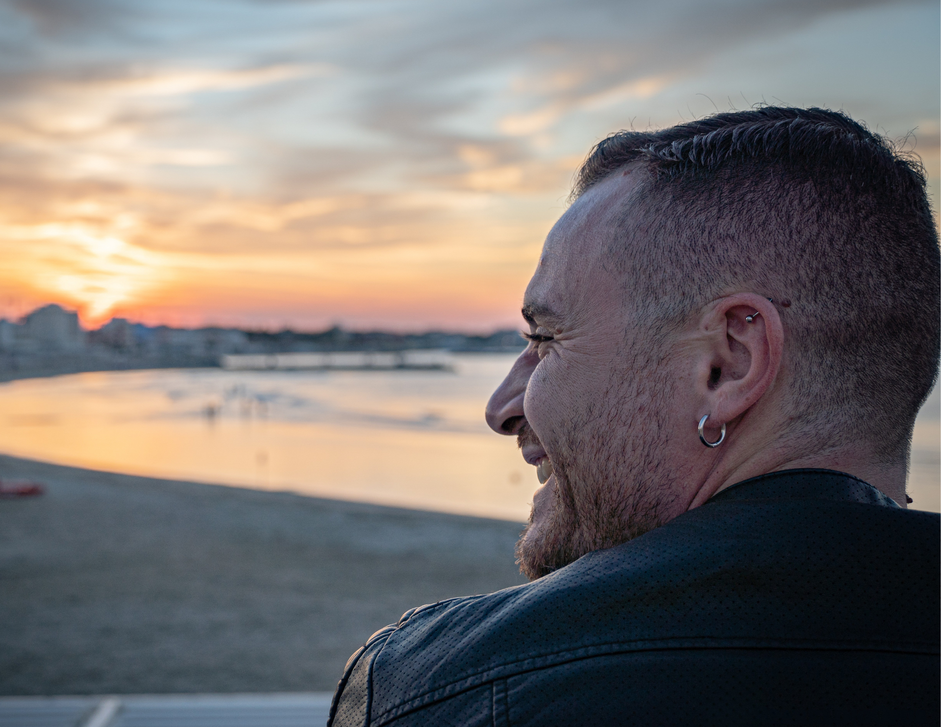 Uomo con la testa rasata che sorride al tramonto su una spiaggia, indossando una giacca di pelle nera.