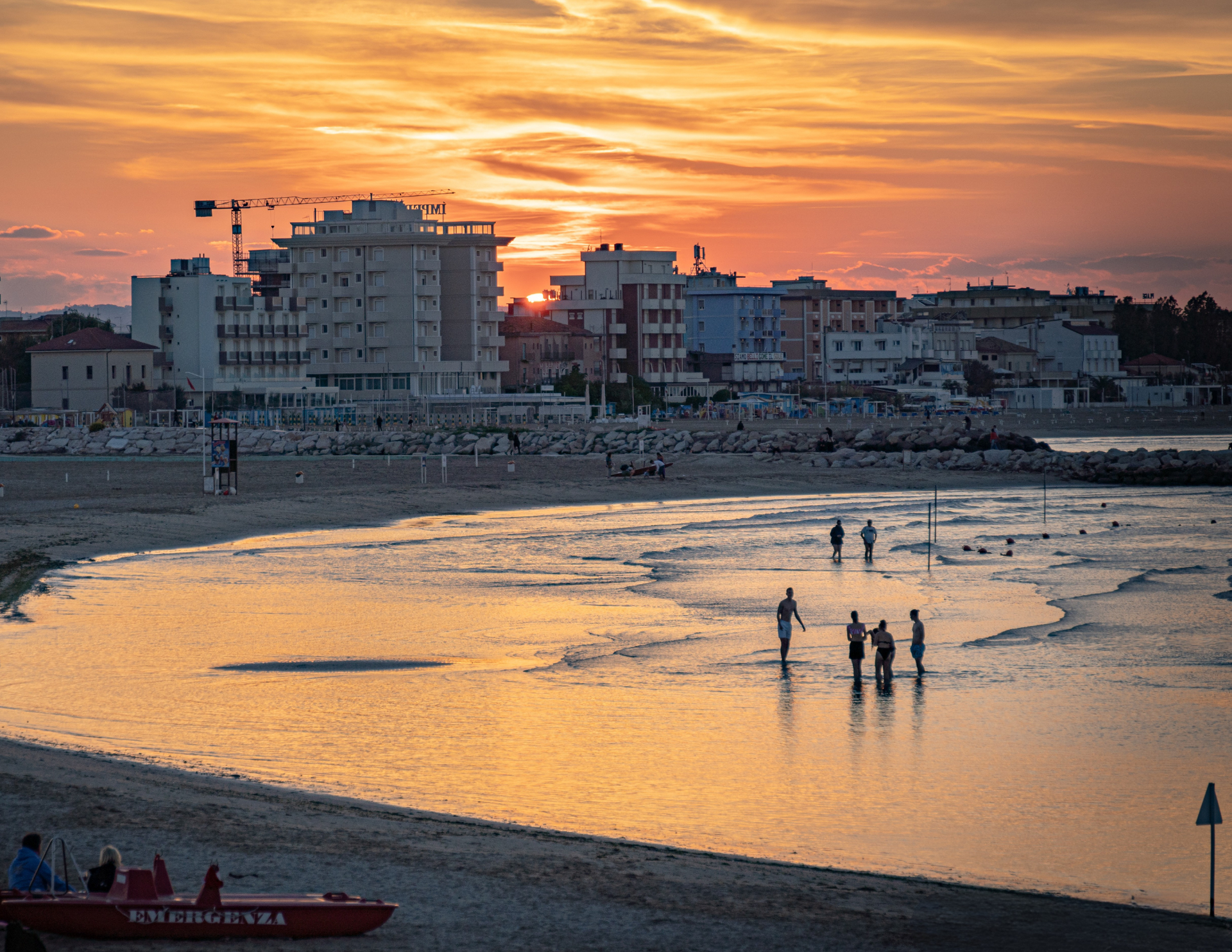 Tramonto su una spiaggia con persone in acqua ed edifici sullo sfondo.
