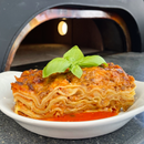 Lasagne in einer weissen Schüssel, mit Basilikum garniert, vor einem Pizzaofen im Restaurant.