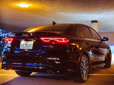 Black Kia Forte sedan parked, taillights illuminated, under an overcast sky.