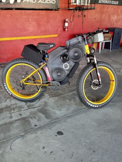 Fat tire bike customized with speakers, yellow rims, and black and gray frame. Red wall backdrop.