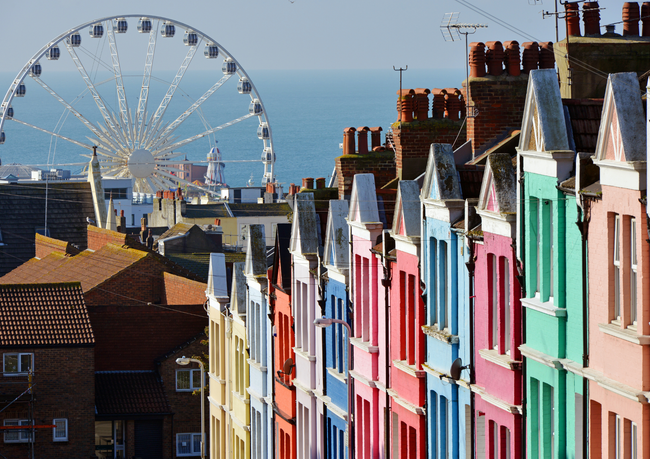 Colorful row houses stand in the foreground, with a large Ferris wheel visible against the sea in the background.