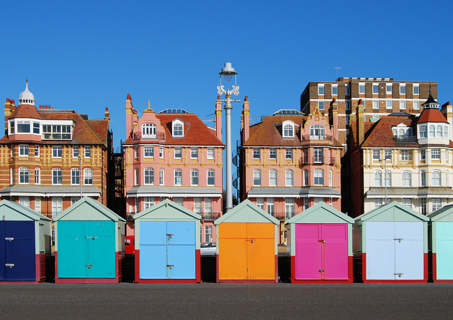 A row of colorful beach huts in front of historic multi-story buildings under a clear blue sky.