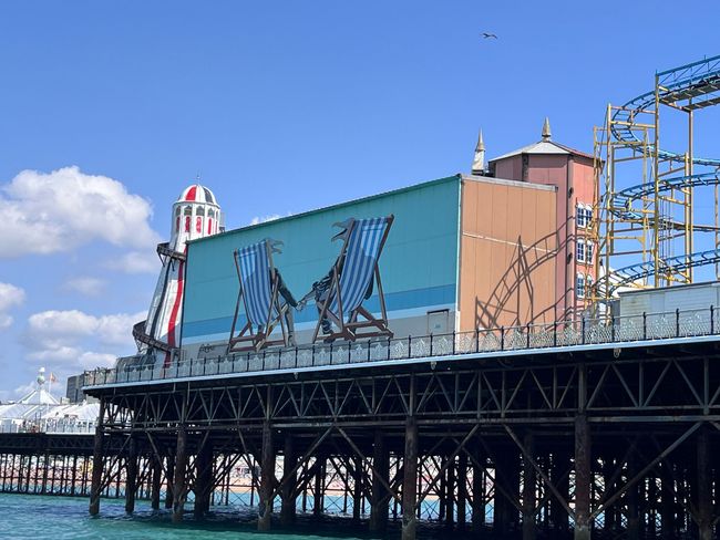 A side view of Brighton Palace Pier, featuring a teal mural of two blue-striped deckchairs against a bright blue sky.