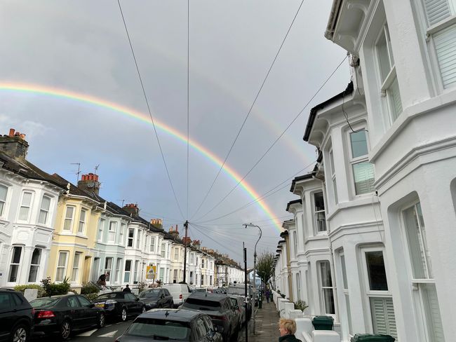 A double rainbow arches over a street lined with row houses and parked cars under a cloudy sky.