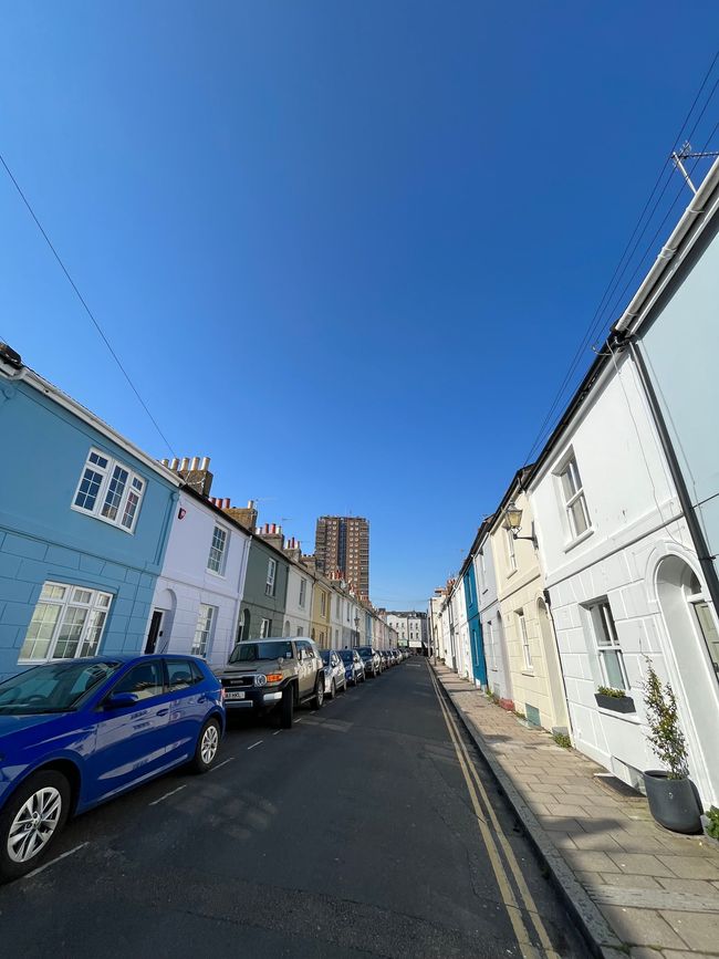 A narrow street lined with colorful houses under a clear blue sky, featuring parked cars along the road.