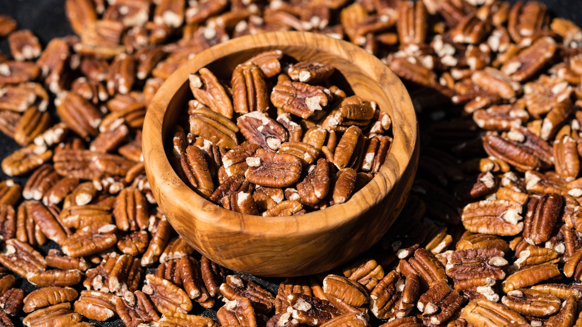 A wooden bowl filled with pecans is sitting on top of a pile of pecans.