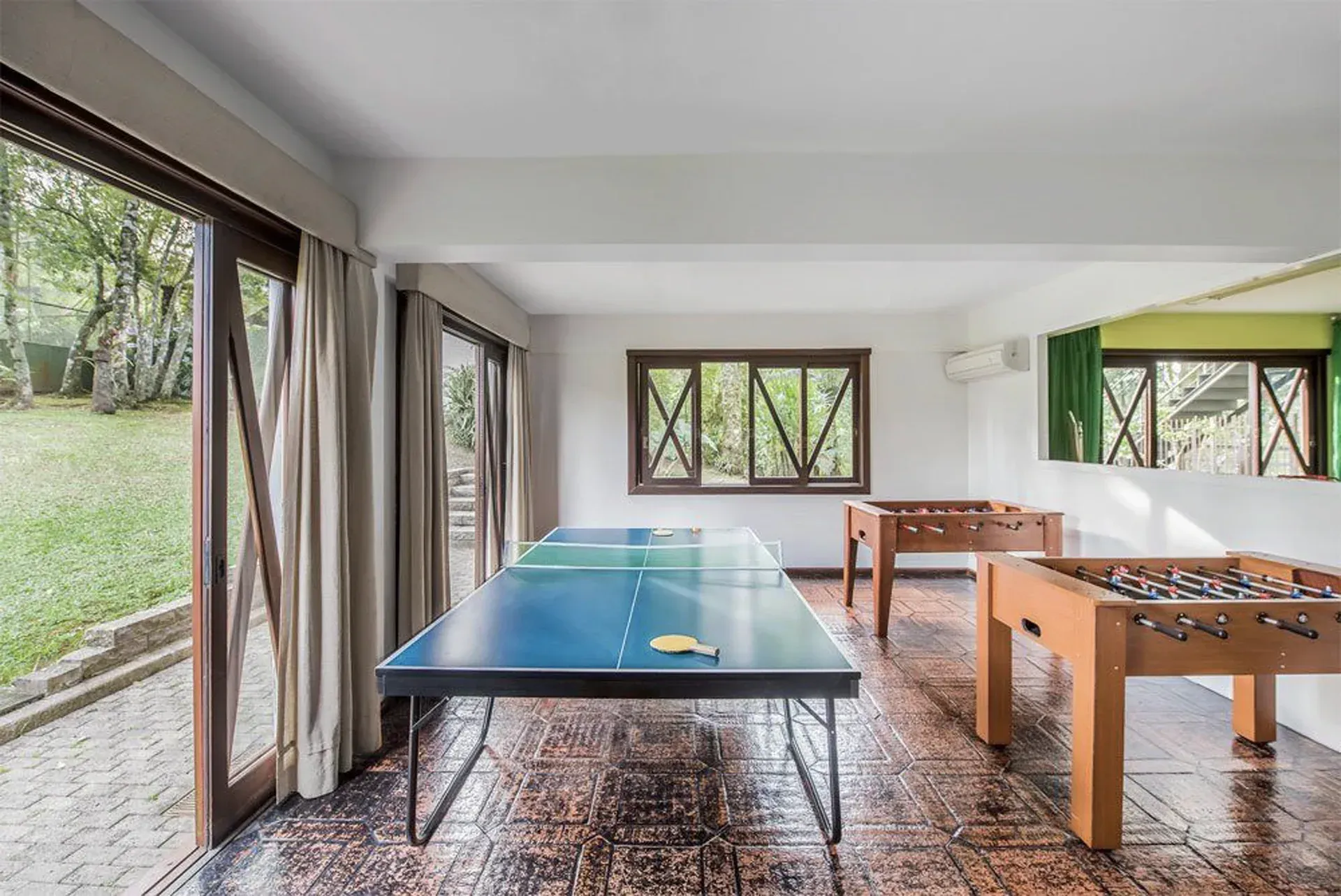 Sala de jogos interna com mesas de pingue-pongue e pebolim, com laterais abertas e vista para a área verde.