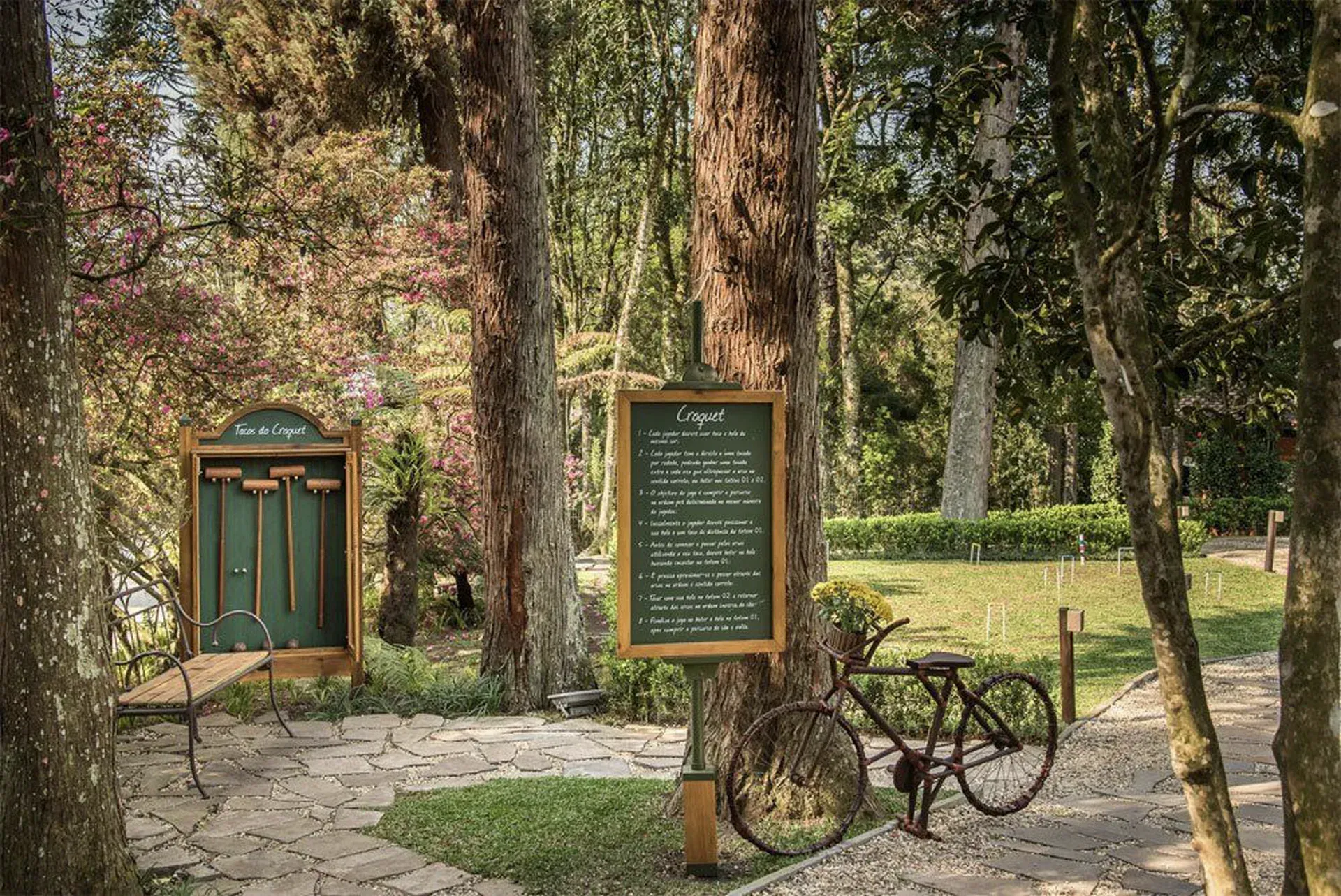 Cena de parque arborizado com placa verde, porta pequena, banco e bicicleta antiga sobre pavimento de pedra.