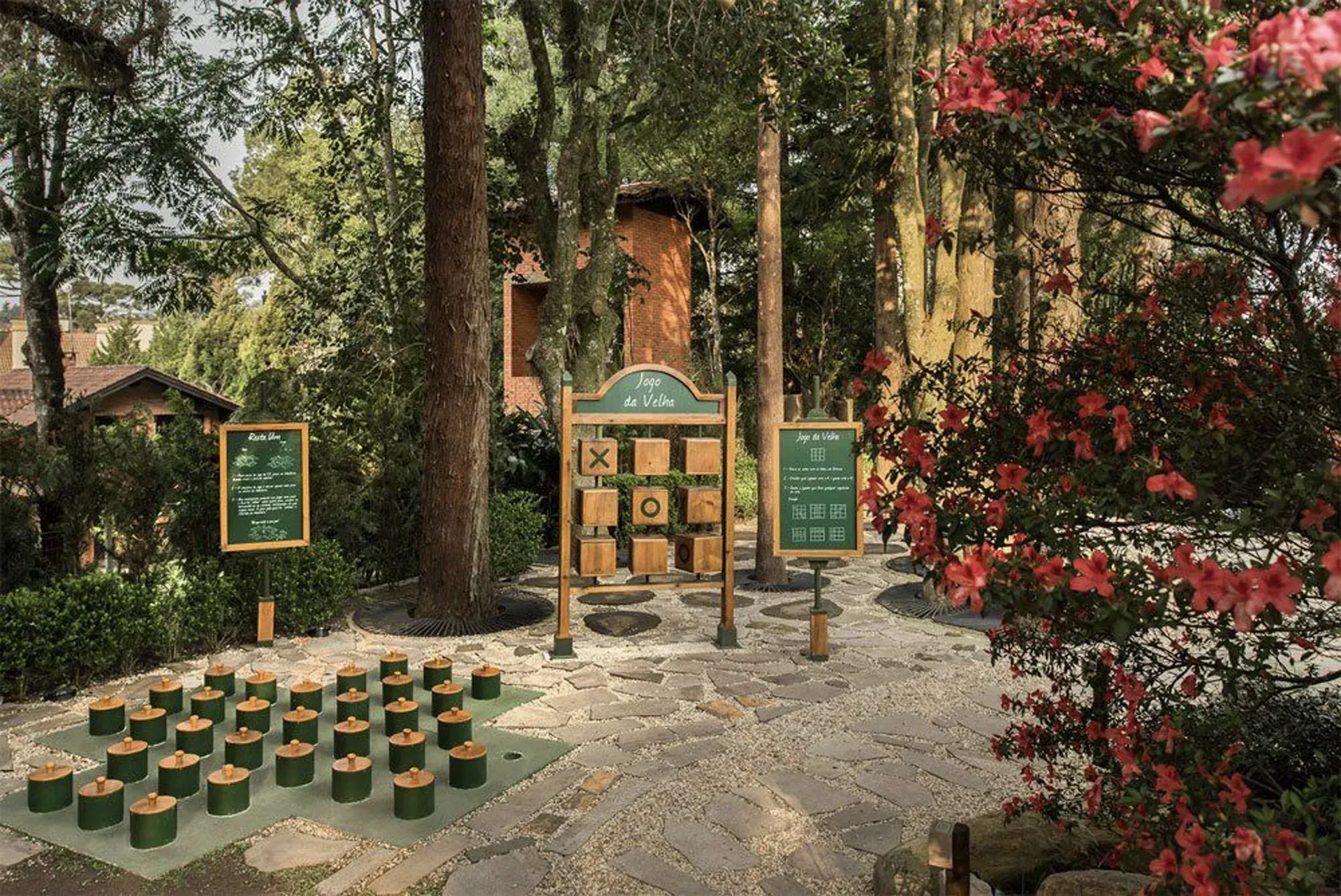 Decoração de jardim ao ar livre com placas de lousa, arranjos de velas e um pequeno suporte de madeira entre árvores e flores vermelhas.