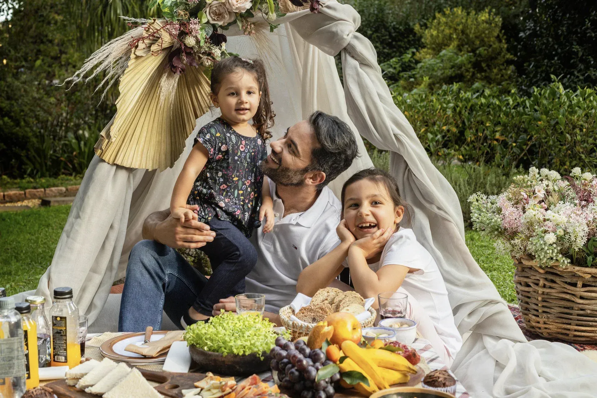Piquenique em família ao ar livre, estendido numa manta, com frutas, flores e uma criança sorridente sob um toldo.