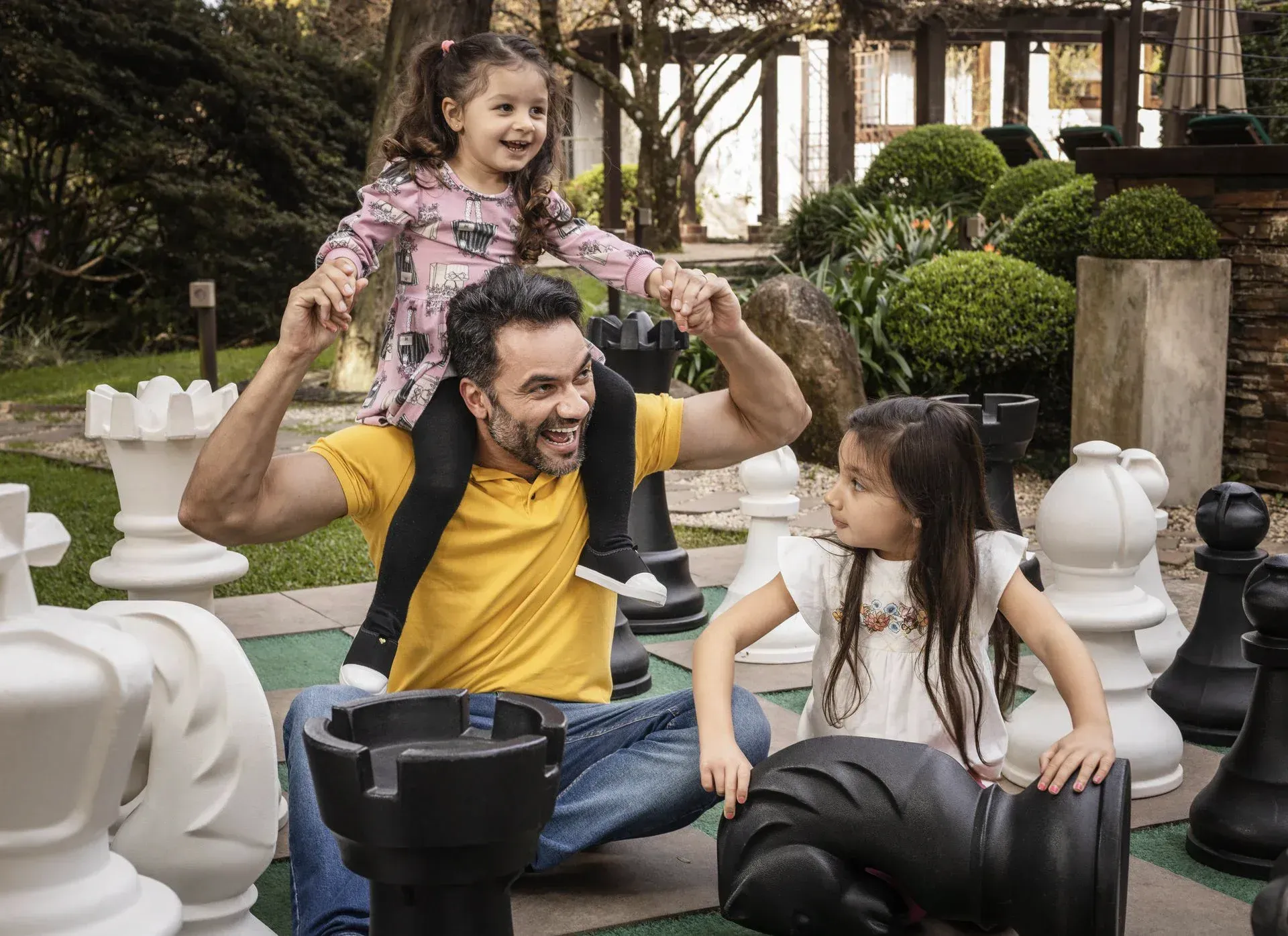 Homem jogando xadrez ao ar livre com duas crianças em peças gigantes, sorrindo em um jardim.