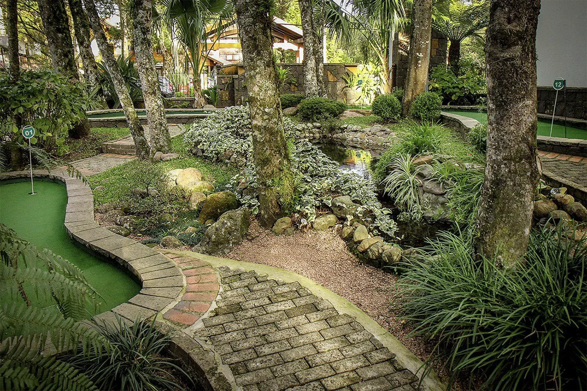 Caminho sinuoso de tijolos que atravessa um jardim exuberante com árvores, arbustos e um pequeno lago verde.