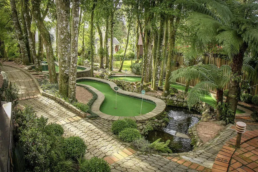 Jardim exuberante com um pequeno lago verde, caminhos de pedra e densas árvores tropicais.