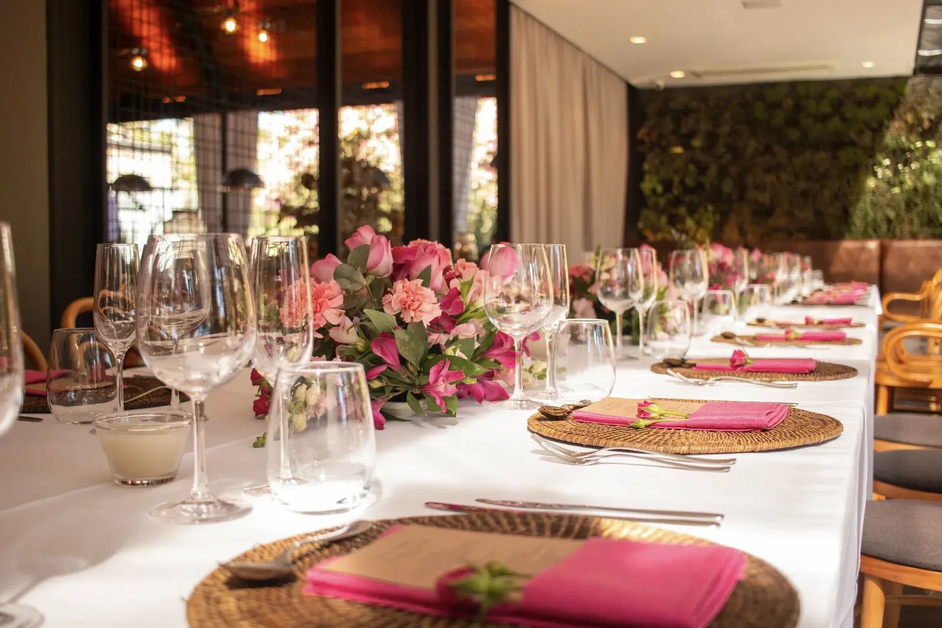 Elegante mesa de banquete com arranjos florais rosa, taças e jogos americanos em tons de rosa e dourado em um restaurante.