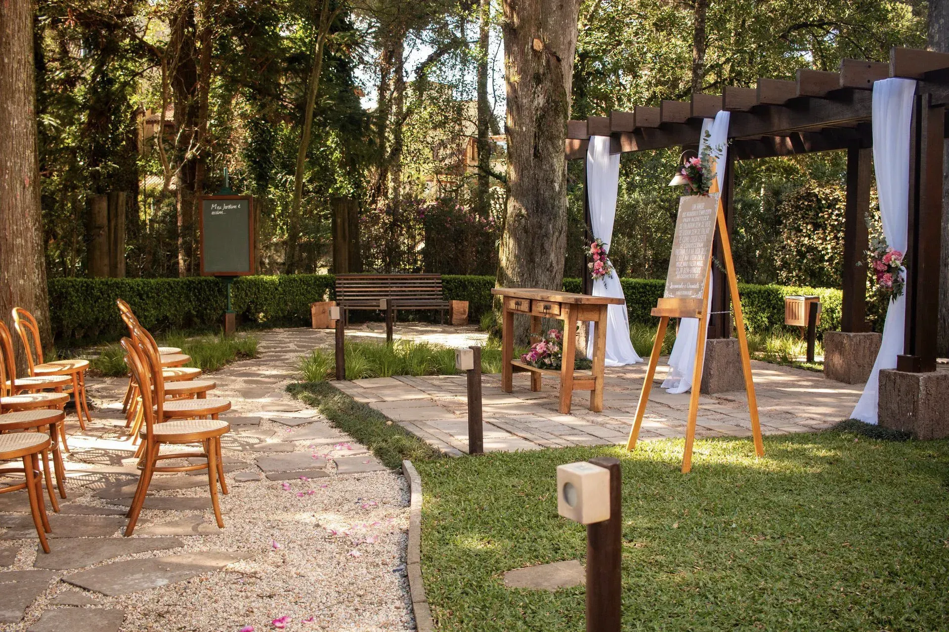 Exposição de arte ao ar livre com cavaletes, cadeiras de madeira e pinturas dispostas ao longo de um caminho de jardim sob árvores.