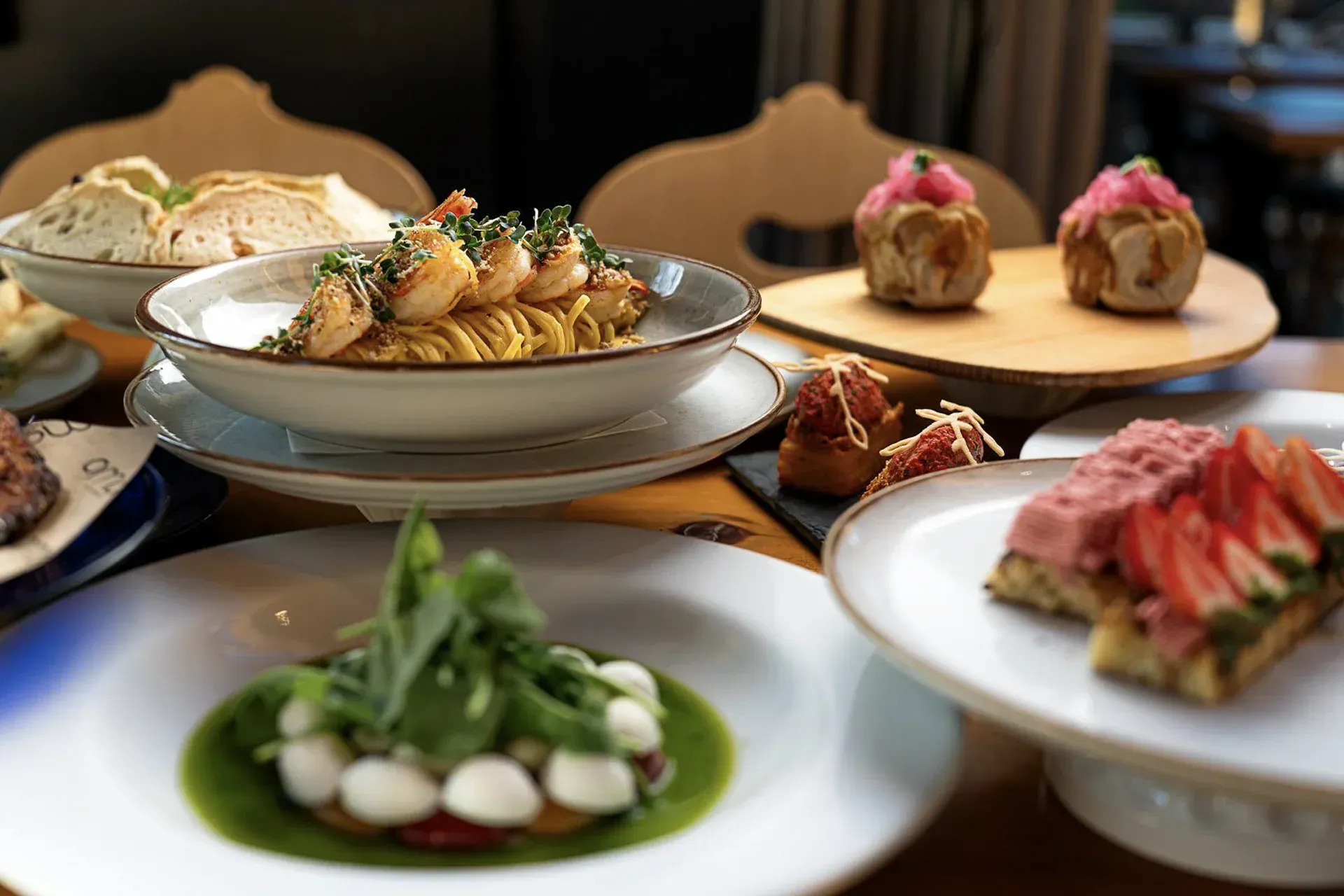 Pratos gourmet variados dispostos em uma mesa de restaurante, incluindo aperitivos e sobremesas com coberturas verdes e rosas.