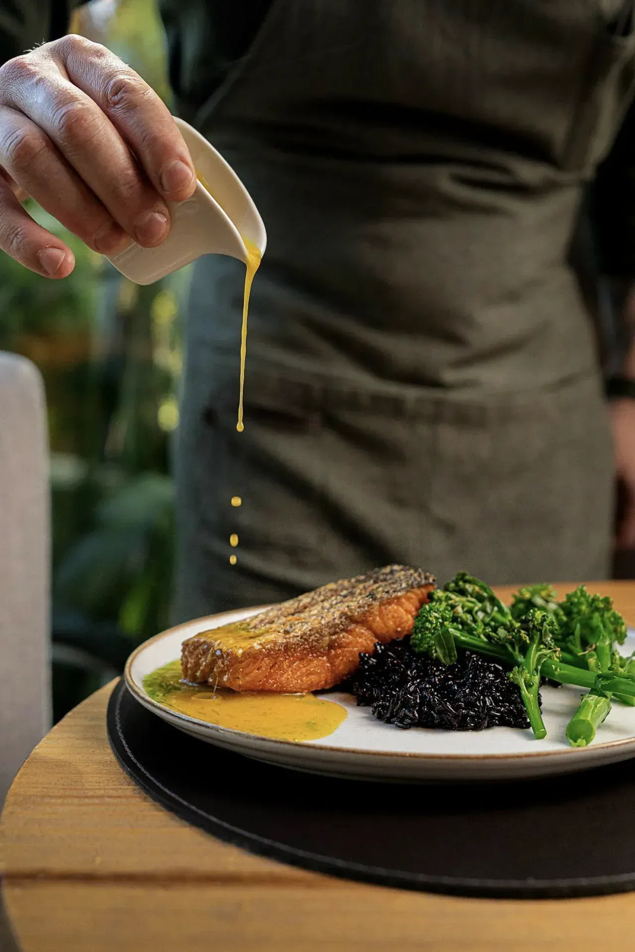 Chef regando molho sobre um prato de peixe com arroz preto e verduras.
