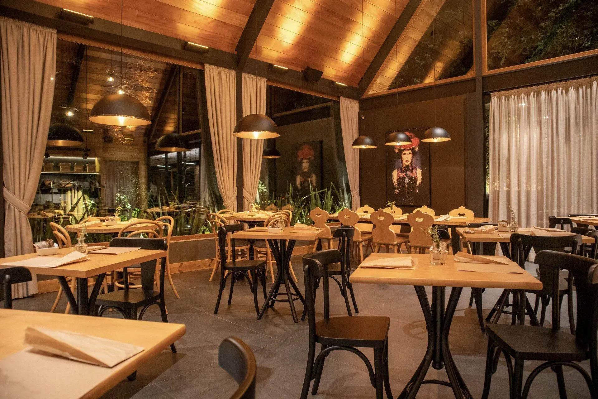 Interior de restaurante rústico com iluminação aconchegante, mesas de madeira, cadeiras pretas e luminárias pendentes.