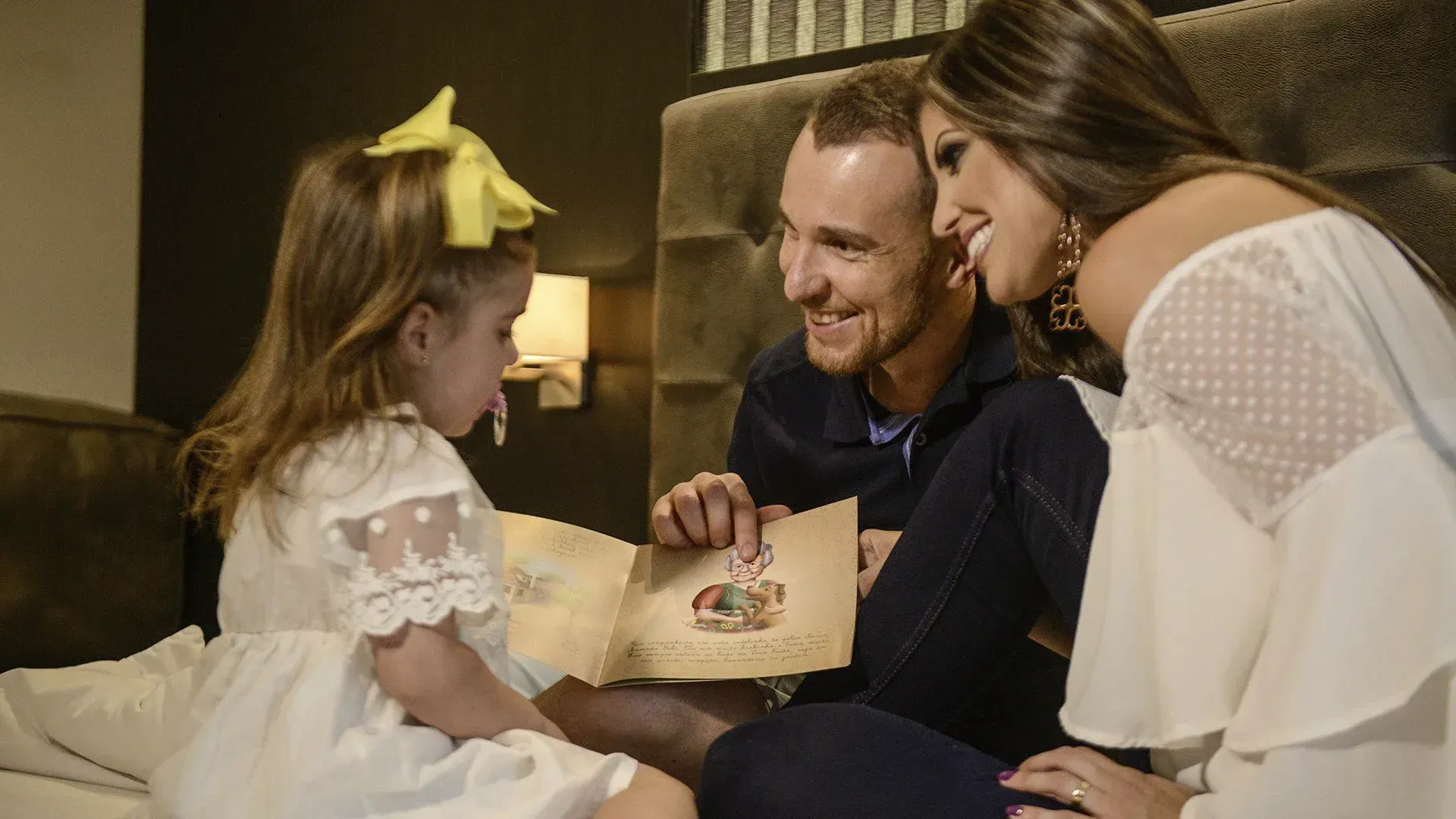 Família lendo um livro de histórias na cama, sorrindo juntos com uma criança pequena de vestido branco e laço amarelo.
