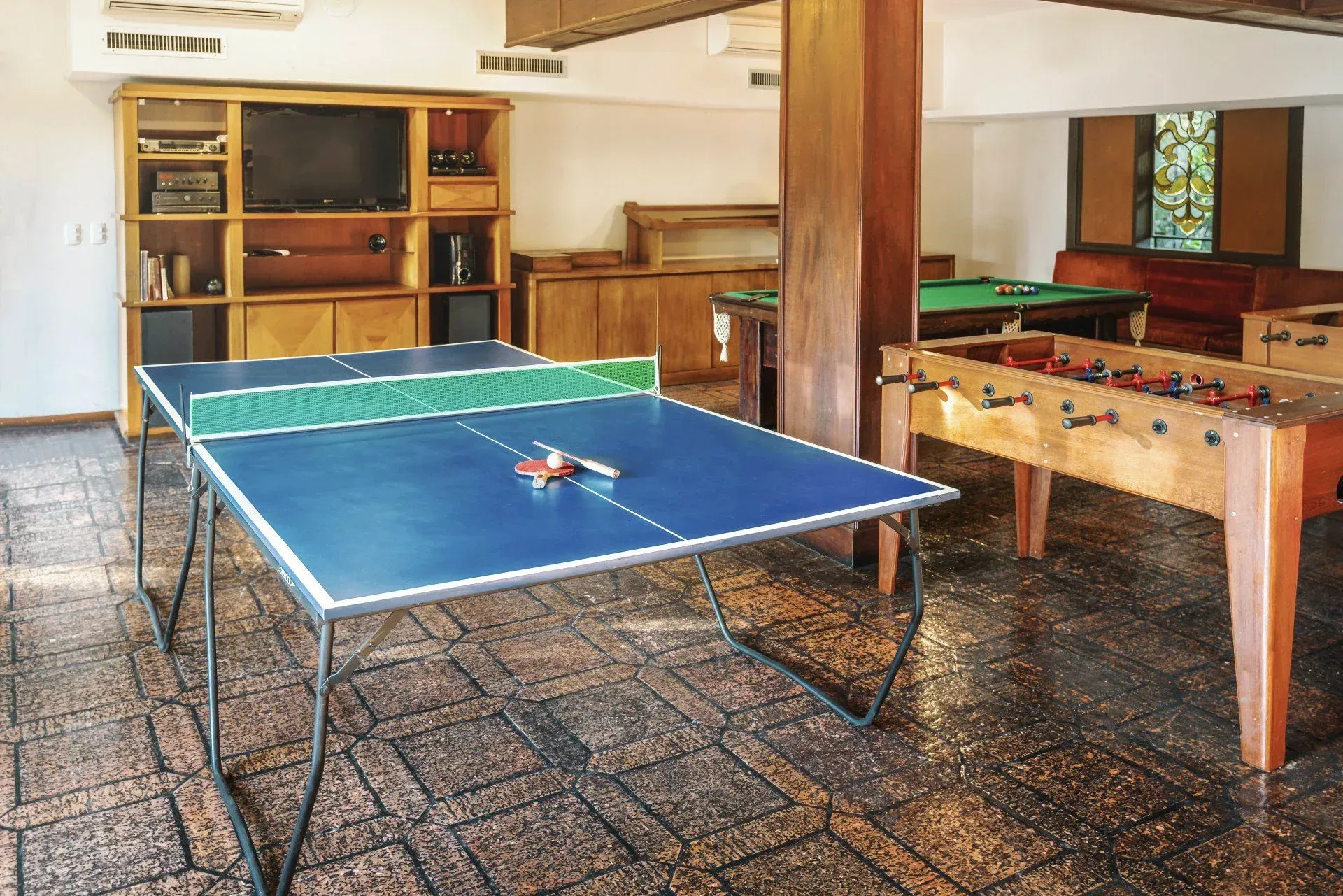 Mesa de pingue-pongue em uma sala de recreação com uma mesa de pebolim e prateleiras de madeira atrás dela.