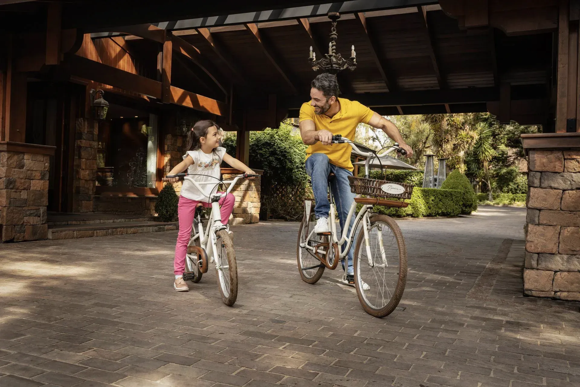 Adulto e criança andando de bicicleta sob um pavilhão de madeira em uma trilha pavimentada.