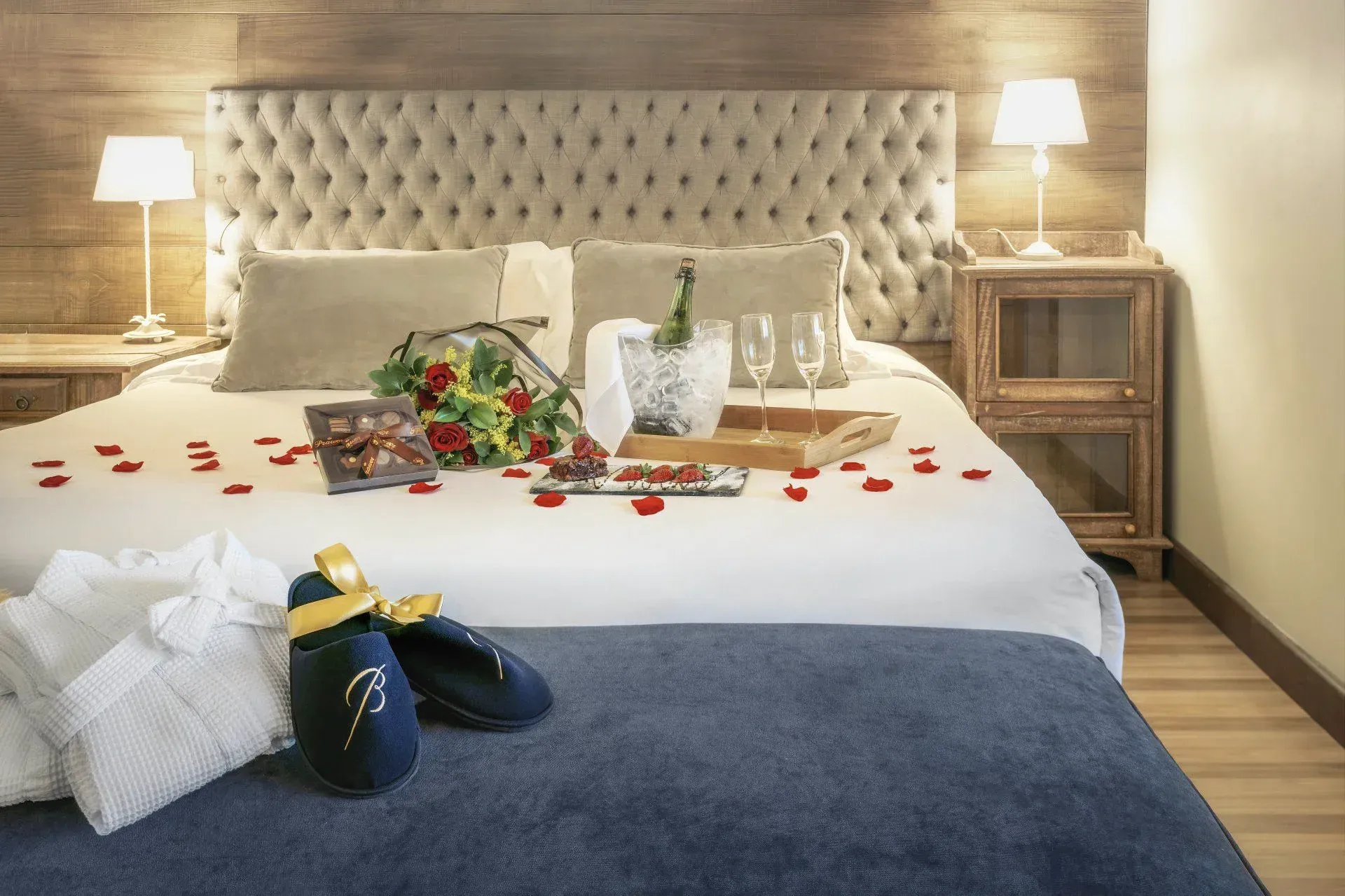 Quarto romântico com pétalas de rosa, champanhe, flores e velas sobre uma cama impecavelmente arrumada.