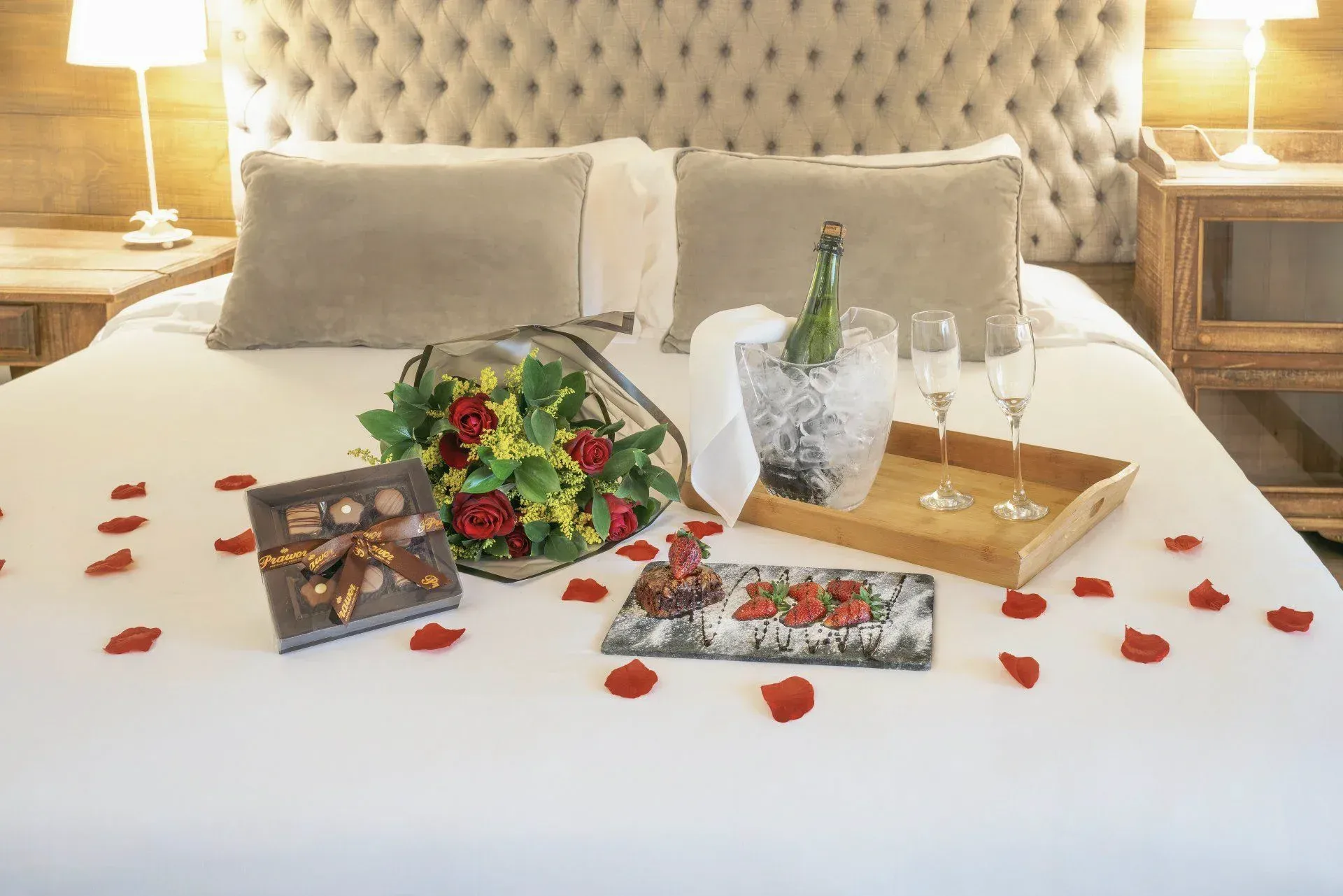 Cama de hotel romântica com pétalas de rosa, flores, chocolate, velas e champanhe sobre um edredom branco.