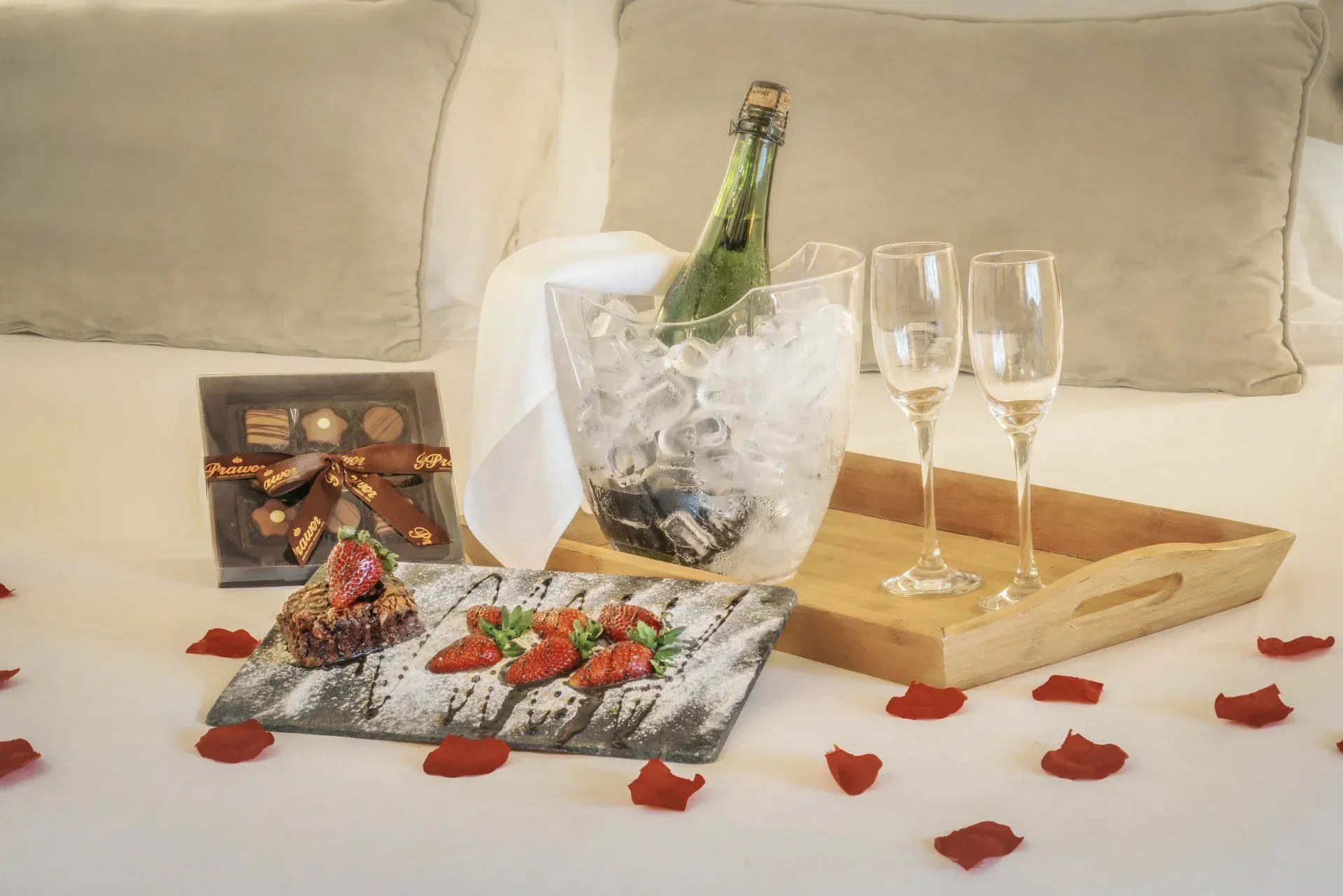 Garrafa de champanhe em balde de gelo com duas taças, chocolates e pétalas de rosa em uma bandeja de cabeceira.