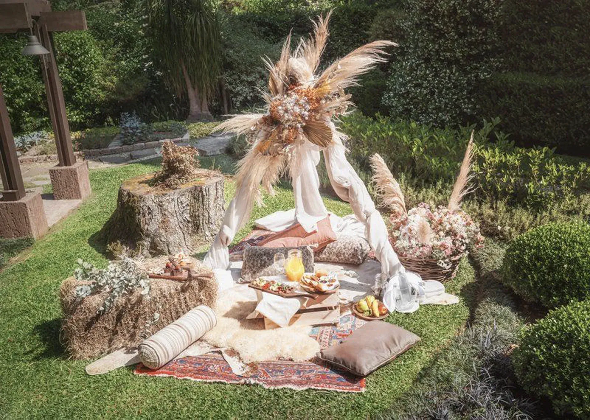 Um altar decorativo ao ar livre com flores, velas, frutas e objetos trançados sobre um tapete em um jardim.