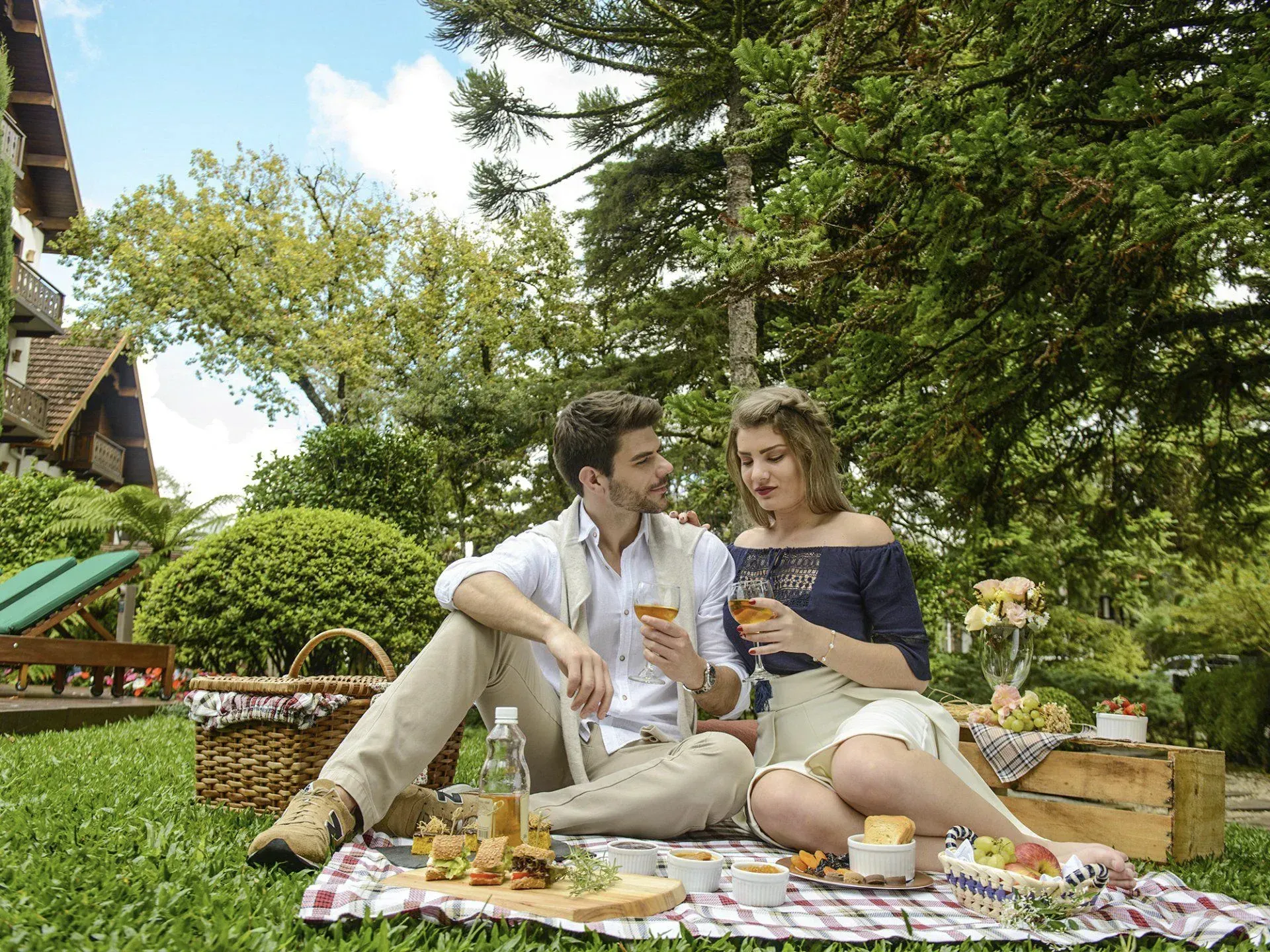 Casal sentado em uma manta de piquenique em um jardim exuberante, compartilhando uma bebida e petiscos sob árvores altas.