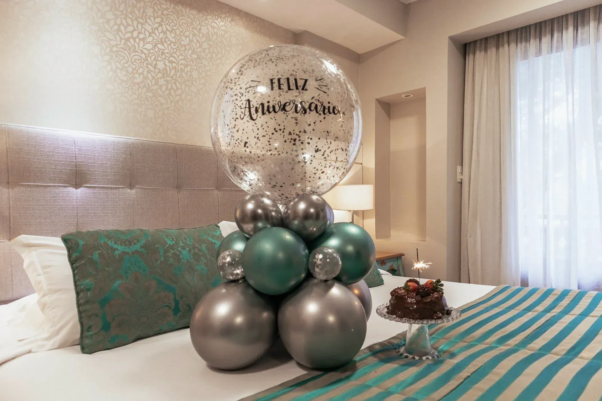 Arranjo de balões de aniversário em uma cama em um quarto com iluminação suave e decoração em tons de azul-petróleo e prata.