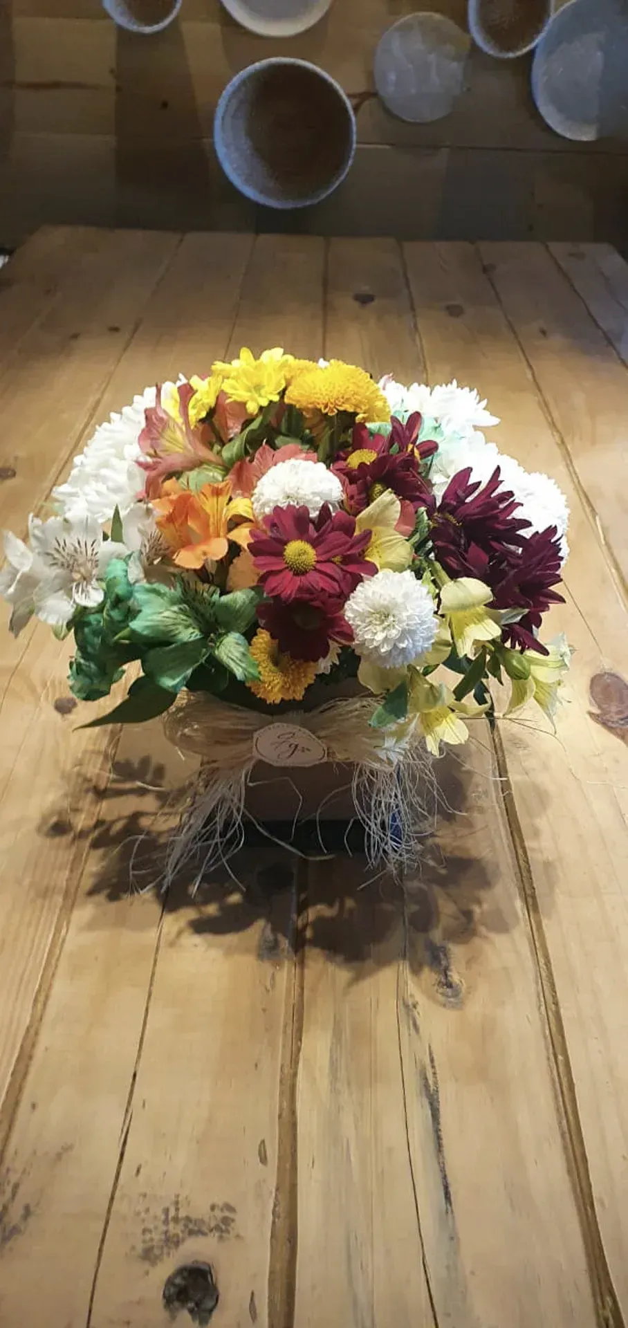 Buquê de flores coloridas em um vaso transparente sobre uma mesa de madeira, com canecas penduradas ao fundo.
