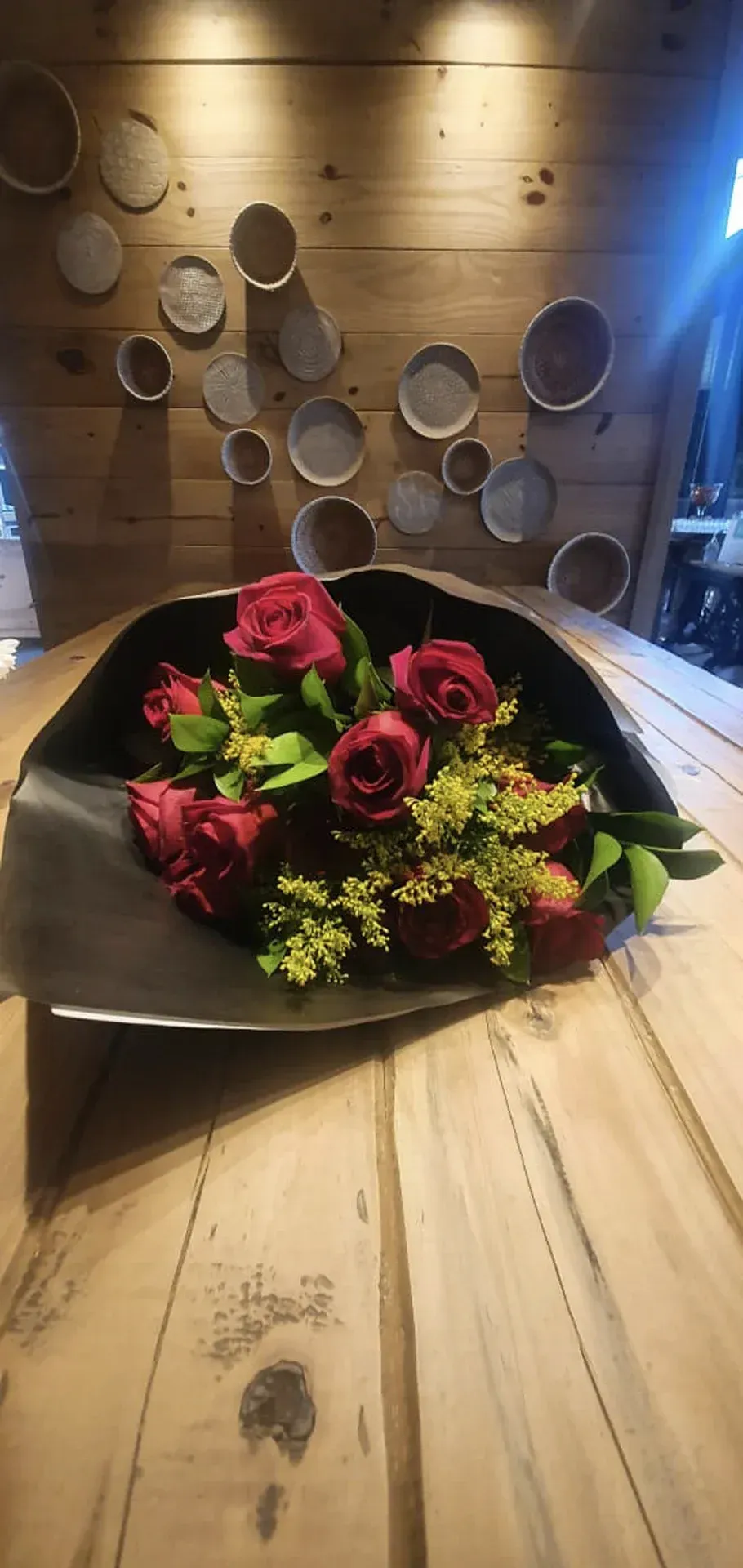 Rosas vermelhas e flores verdes arranjadas em uma tigela preta sobre uma mesa de madeira encostada em uma parede rústica de madeira.