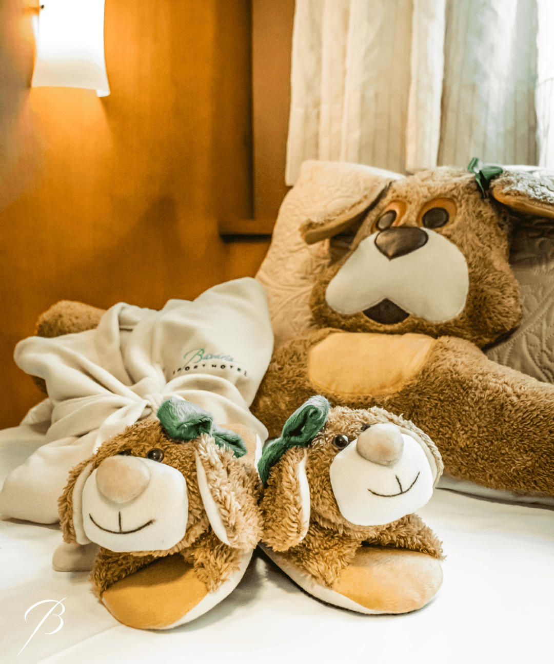 Pantufas de pelúcia em formato de ursinho sobre uma cama ao lado de um urso de pelúcia, em um quarto com iluminação aconchegante.