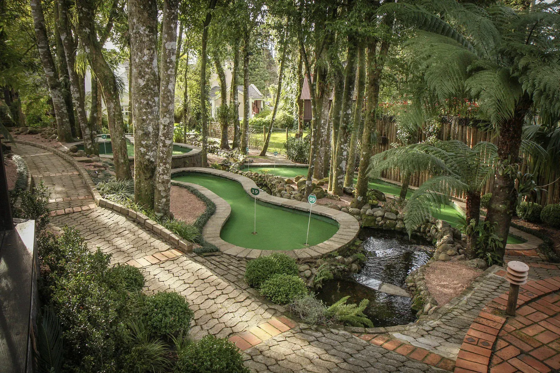 Campo de minigolfe serpenteando por um exuberante jardim arborizado com um pequeno riacho e caminho de tijolos.