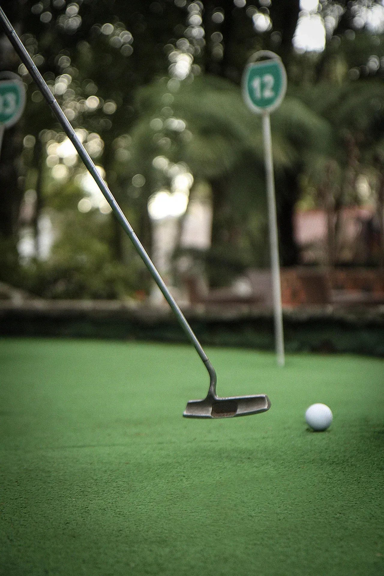 Taco e bola de golfe em um campo de minigolfe verde, com um marcador de buraco ao fundo.