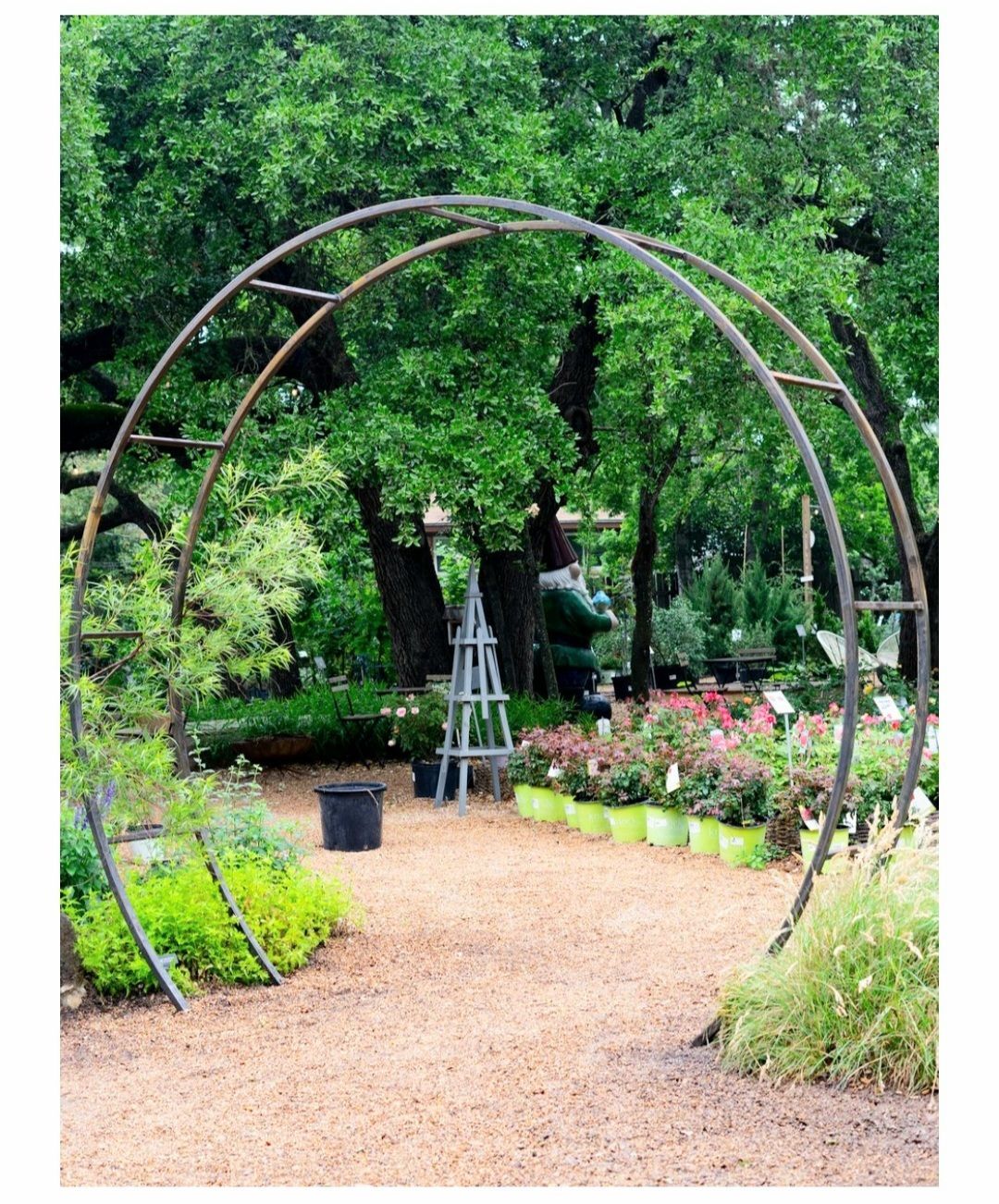 A metal archway in a garden surrounded by trees and potted plants.