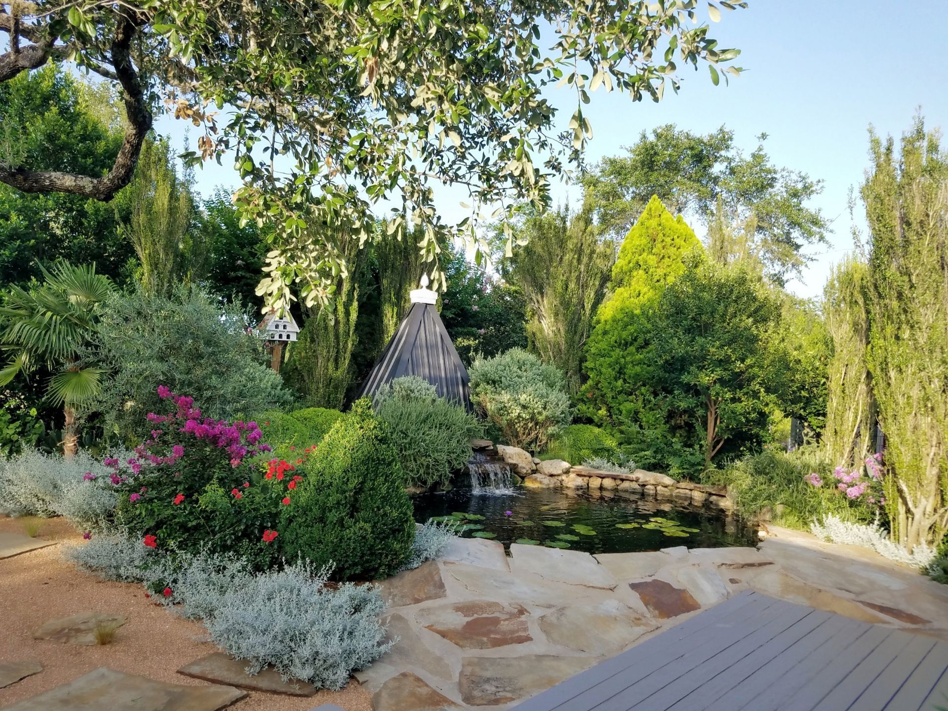 There is a pond in the middle of the garden surrounded by trees and flowers.