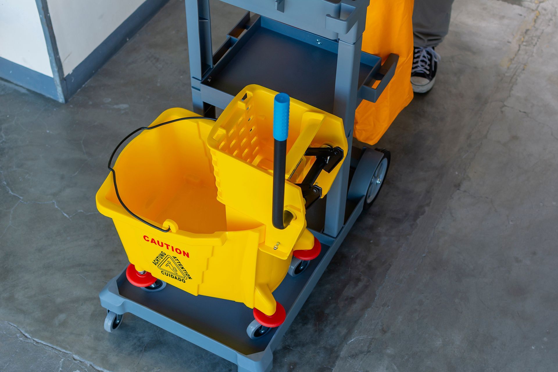 A person is standing next to a yellow mop and bucket on a cart.