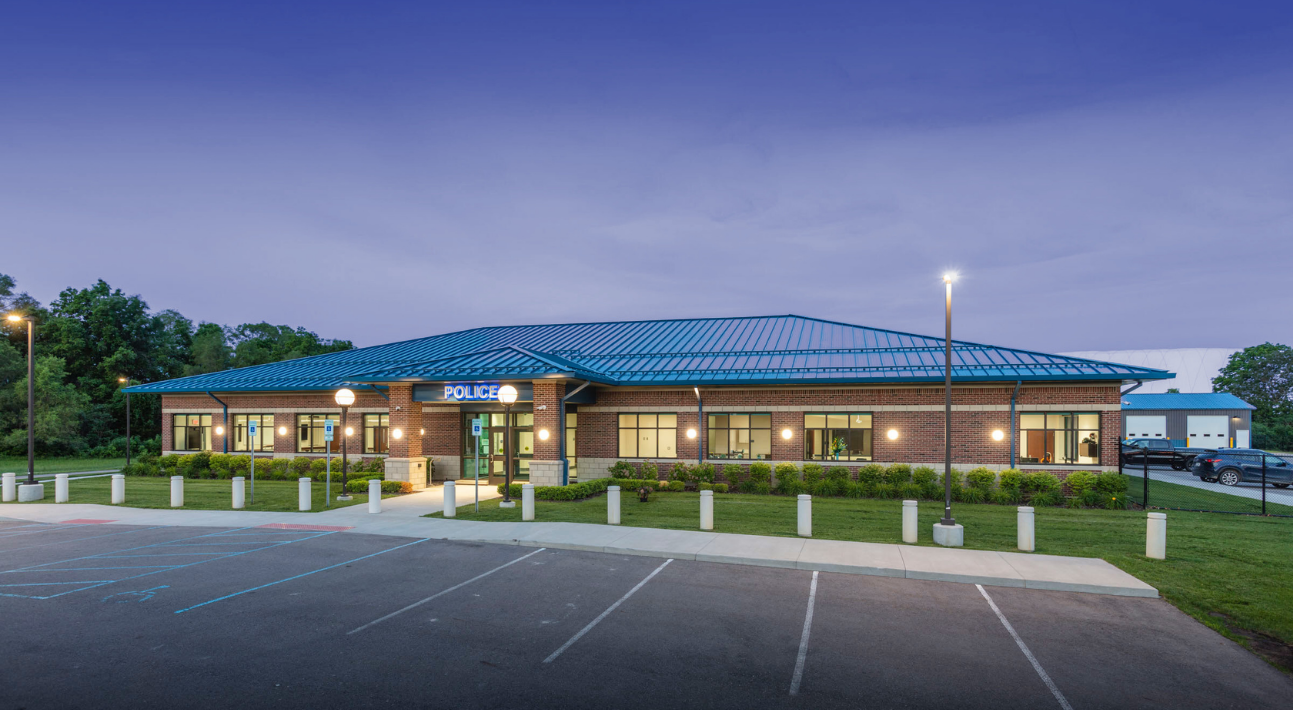 Police station, brick building, blue roof, parked cars in front, trees in the background.