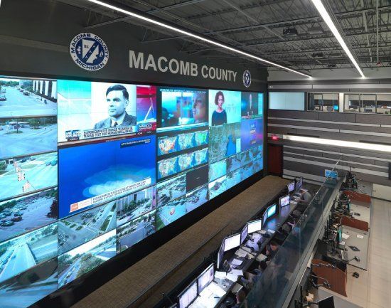 Control room in Macomb County with multiple screens displaying live video feeds and news broadcasts.