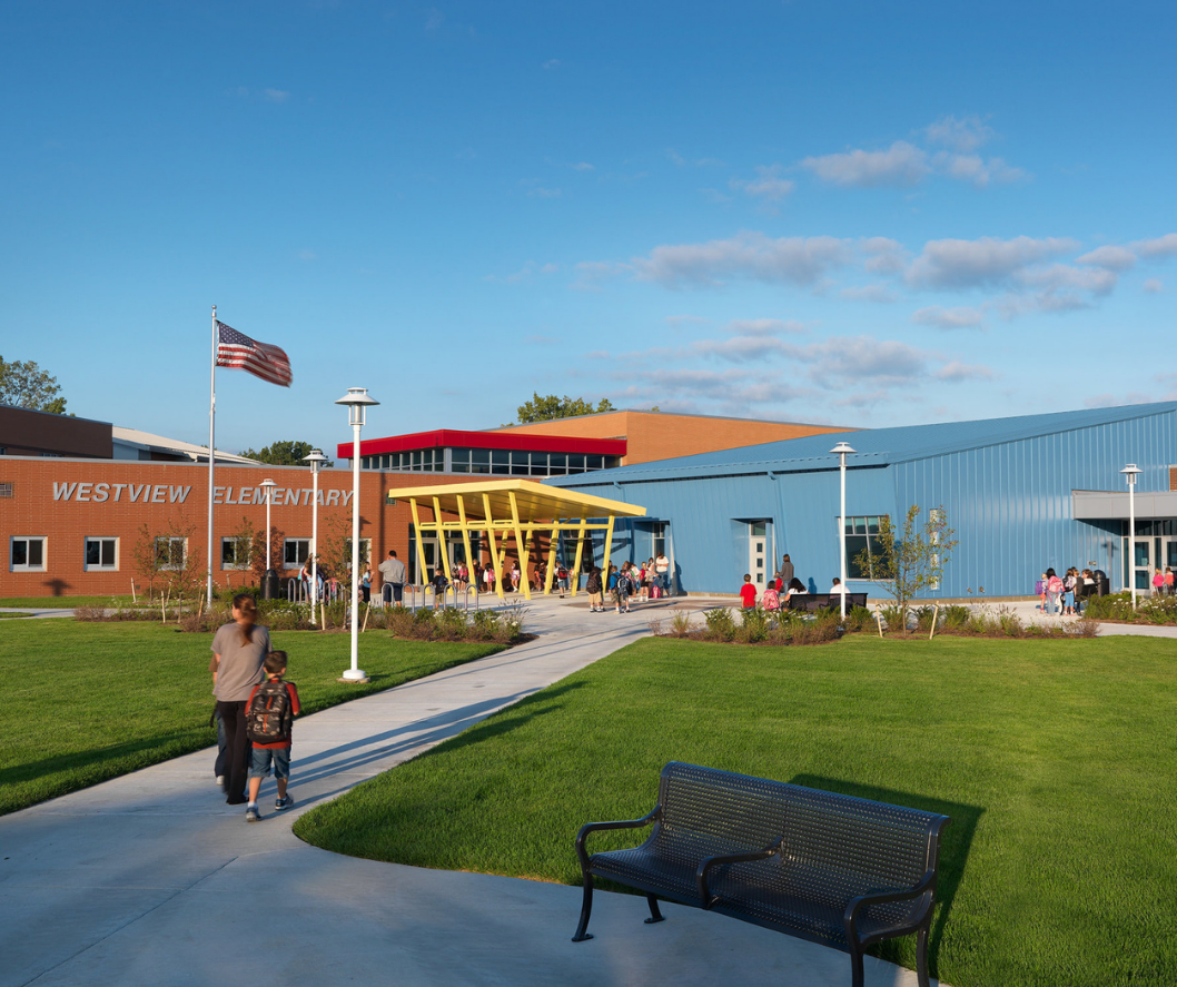 School exterior with flag, people walking. Red brick building with blue and yellow accents, sunny day.