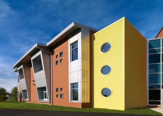 Modern building with orange, yellow, and silver panels, blue sky. Three circular windows on yellow wall.