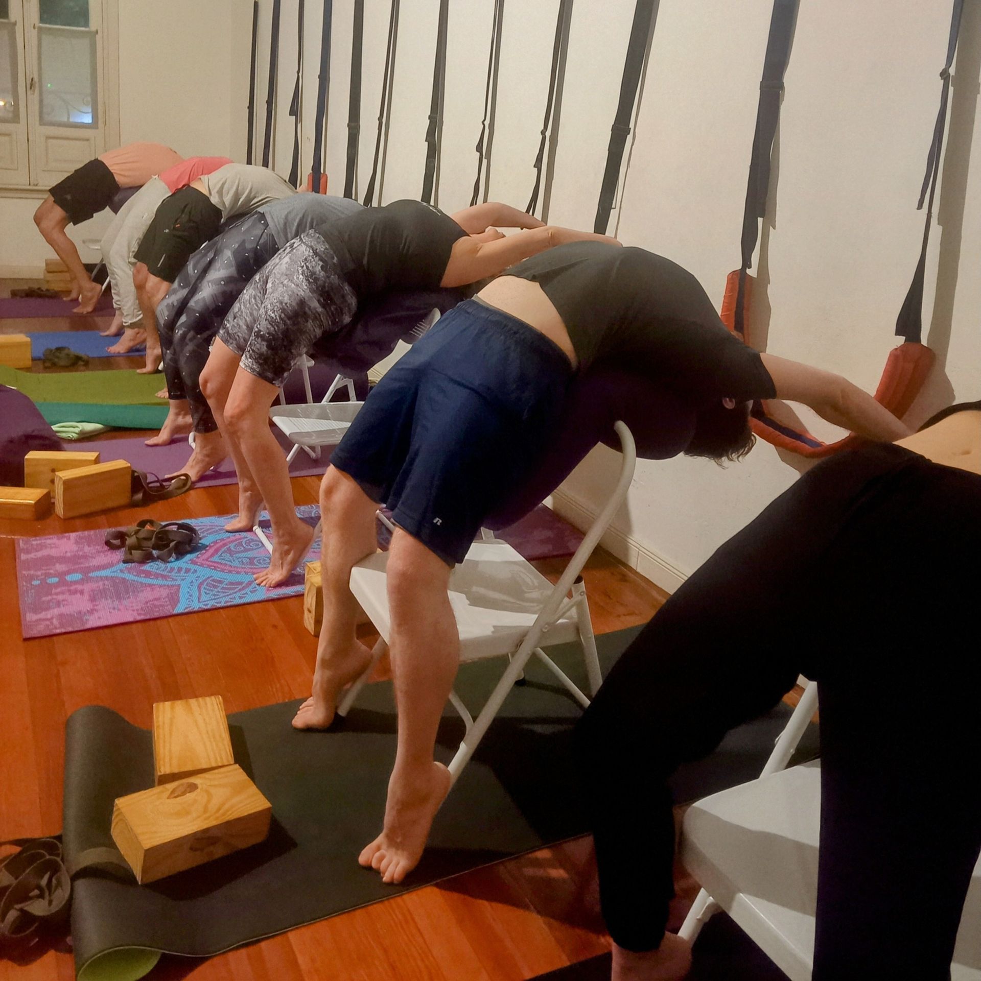 Personas haciendo posturas de yoga con sillas y cuerdas de pared en un estudio con piso de madera.