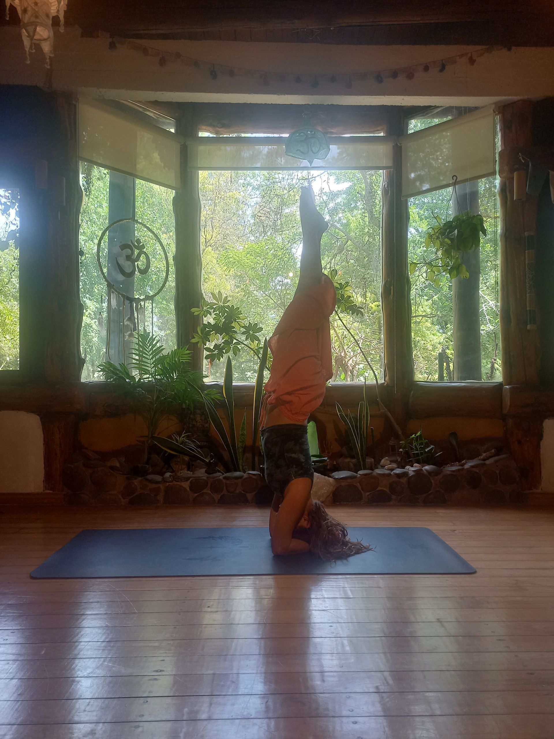 Persona en pose de parada de manos sobre una colchoneta de yoga azul, en una habitación con suelo de madera y grandes ventanales.